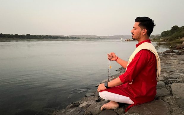 A priest meditating near a serene riverside embodies the essence of spiritual connection and inner peace. Sitting in a calm and reflective posture, the priest e